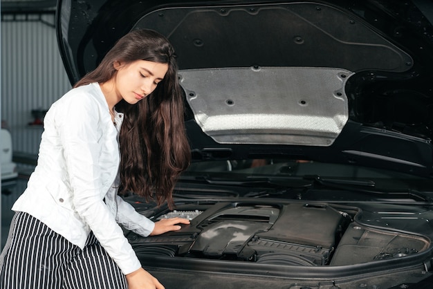 Giovane donna in piedi davanti all'auto con il cofano aperto in garage guardando sotto il cofano dell'auto