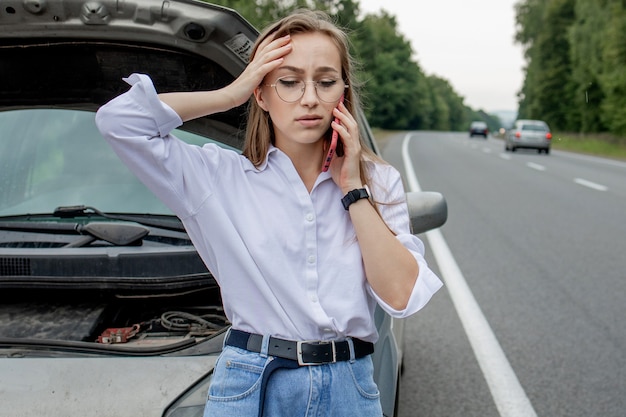 Giovane donna in piedi vicino a un'auto in panne con il cofano sollevato che ha problemi con il suo veicolo.