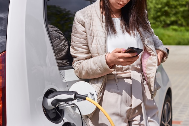 Giovane donna in piedi vicino all'auto elettrica con il cellulare in mano e in attesa di ricarica della batteria dell'automobile.