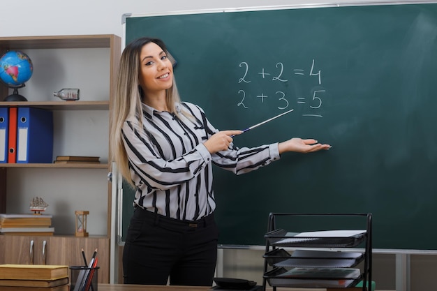 giovane donna in piedi vicino alla lavagna in classe spiegando la lezione tenendo il puntatore che si presenta con il braccio della mano sorridente fiducioso