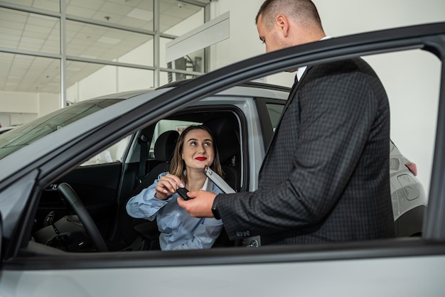 Giovane donna in piedi vicino alla nuova auto moderna presso la concessionaria e scegliendo la migliore auto con il venditore