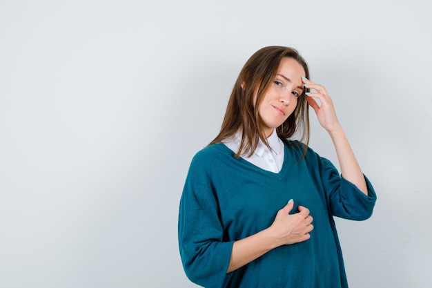 Giovane donna in posa mentre si tocca la fronte con le dita in camicia bianca, maglione e sembra sensata. vista frontale.