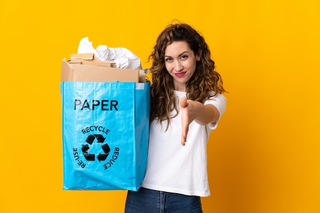 Giovane donna in possesso di un sacchetto di riciclaggio pieno di carta da riciclare isolato su sfondo giallo si stringono la mano per chiudere un buon affare