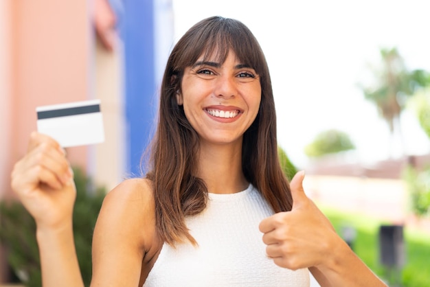 Giovane donna in possesso di una carta di credito all'aperto con i pollici in su perché è successo qualcosa di buono