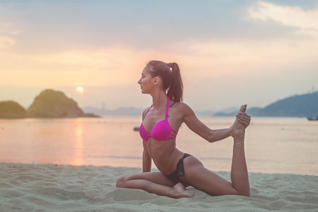 Giovane donna in reggiseno rosa e mutandine nere seduta sulla spiaggia allungando le gambe durante il tramonto in mare Ragazza fitness facendo esercizi in riva al mare