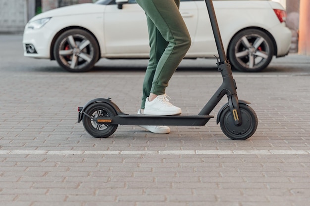 Giovane donna in scarpe da ginnastica bianche è in piedi su uno scooter elettrico davanti all'auto bianca