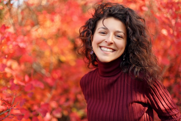 Giovane donna in sweather rosso che posa sulla macchina fotografica in cespugli autumnn rossi