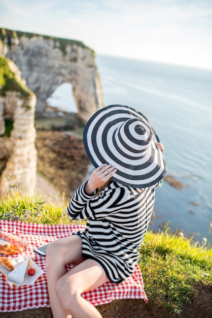 Giovane donna in testa a strisce e maglione avente un picnic con vino, formaggio e pane seduto sulla costa rocciosa in France
