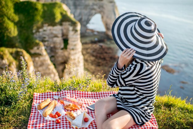 Giovane donna in testa a strisce e maglione avente un picnic con vino, formaggio e pane seduto sulla costa rocciosa in France