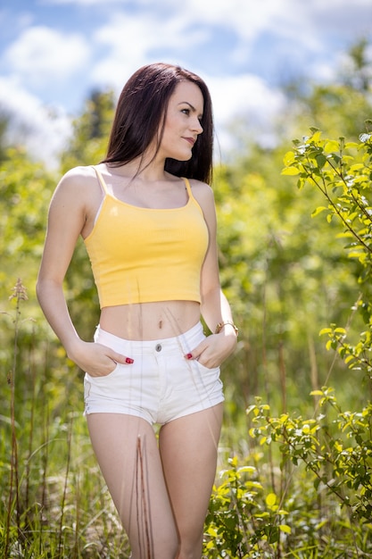 Giovane donna in top giallo e pantaloncini bianchi in piedi nella natura in estate