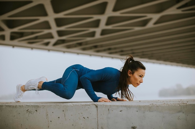 Giovane donna in tuta blu che fa flessioni sotto il ponte nell'ambiente urbano