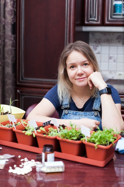 Giovane donna in tuta in denim in cucina prendersi cura di piantine di erbe