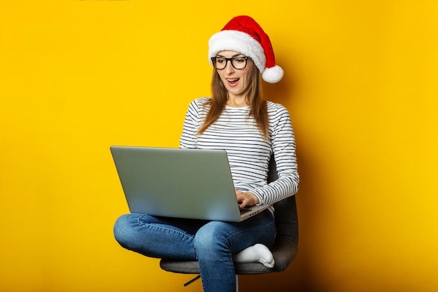 Giovane donna in un cappello da Babbo Natale con una faccia sorpresa si siede su una sedia e tiene un computer portatile