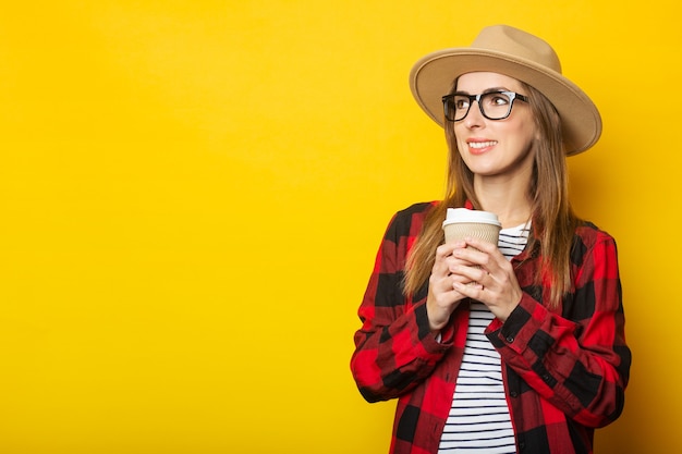 Giovane donna in un cappello e una camicia a quadri tiene un bicchiere di carta con caffè su una superficie gialla