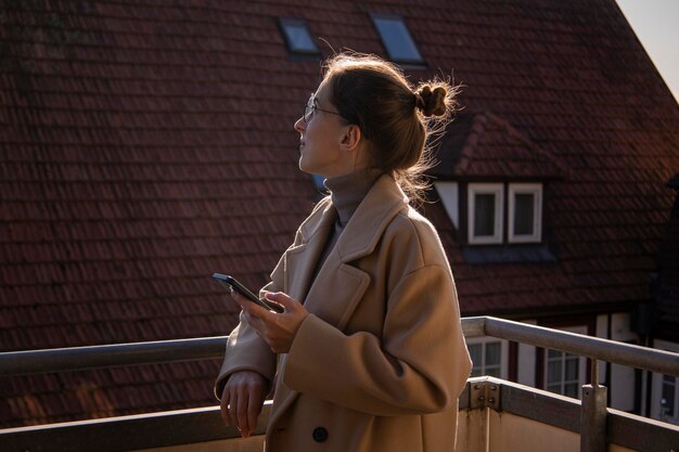 Giovane donna in un cappotto con un telefono in piedi sulla terrazza