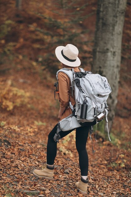 Giovane donna in un elegante cappello e borsa da viaggio sulle spalle, guardando intorno alla foresta di autunno affascinante