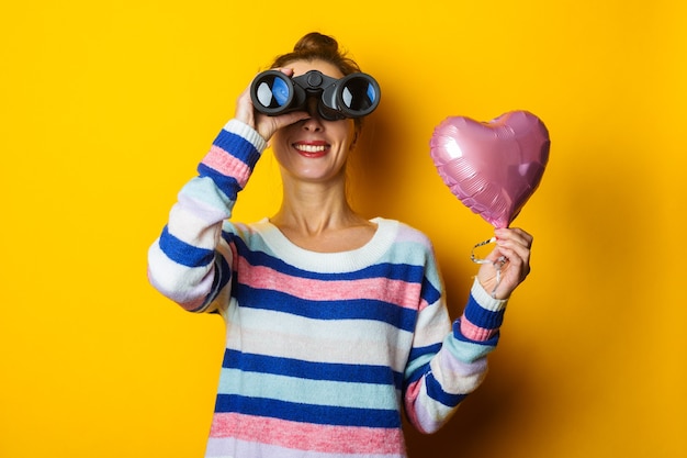 Giovane donna in un maglione tiene un cuore di mongolfiera e guarda attraverso il binocolo su uno sfondo giallo. Composizione di San Valentino.