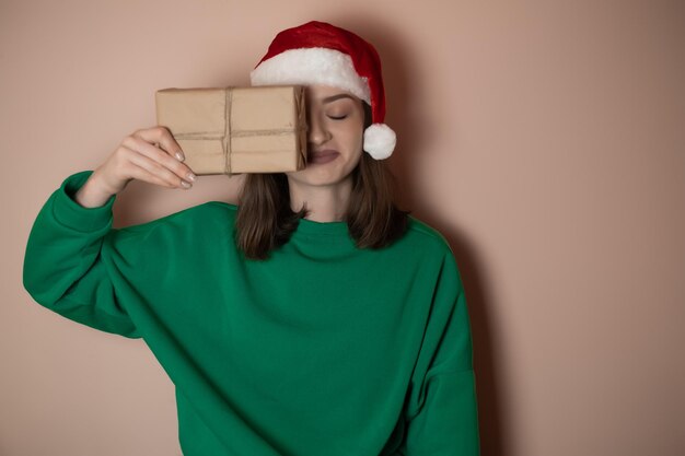 Giovane donna in un maglione verde e in un cappello di Capodanno tiene una confezione regalo su un semplice sfondo verde Concetto di vacanza Happy New Year 2023 chrome key buon natale