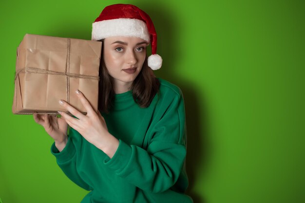 Giovane donna in un maglione verde e in un cappello di Capodanno tiene una confezione regalo su un semplice sfondo verde Concetto di vacanza Happy New Year 2023 chrome key buon natale