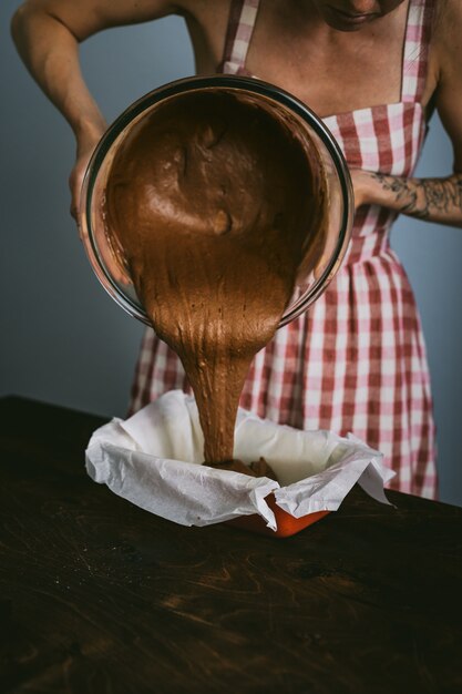 giovane donna in un vestito a quadri rosso e bianco cuocere una torta, versando la pastella di cioccolato in una teglia