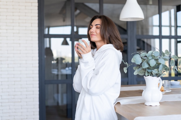 Giovane donna in una calda felpa bianca a casa in cucina, inizia la sua giornata con una tazza di caffè