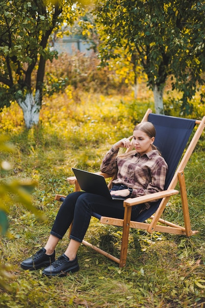 Giovane donna in una camicia a scacchi seduta su una sedia all'aperto in un giardino e che lavora su un computer portatile lavoro a distanza libera professionista femminile