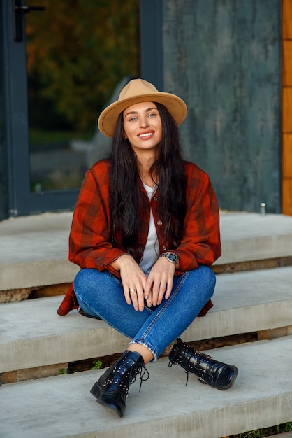 Giovane donna in una camicia rossa alla moda cappello marrone e pantaloni di jeans si siede su una scala, vicino a casa sua.