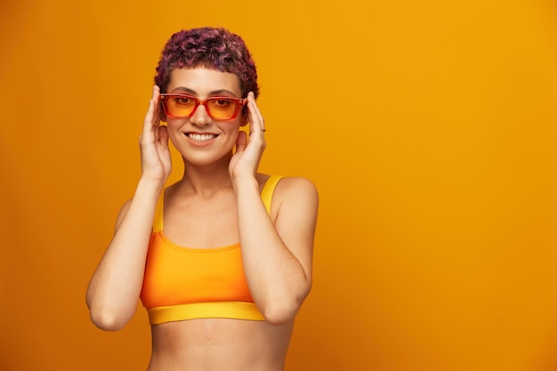 Giovane donna in una tuta fitness giallo brillante con una pancia aperta e occhiali da sole su uno sfondo arancione sorridente