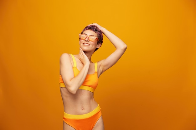 Giovane donna in una tuta fitness giallo brillante con una pancia aperta e occhiali da sole su uno sfondo arancione sorridente
