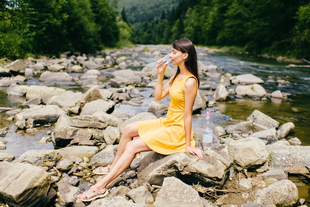 Giovane donna in vestito giallo che si siede sulle pietre nel fiume e nell'acqua potabile