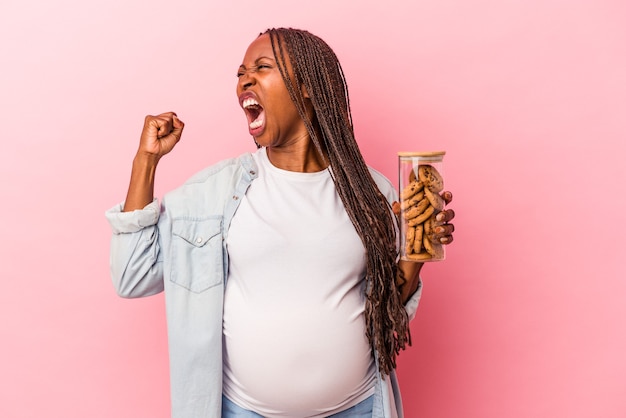 Giovane donna incinta afroamericana che tiene i biscotti isolati su fondo rosa che alza il pugno dopo una vittoria, concetto del vincitore.