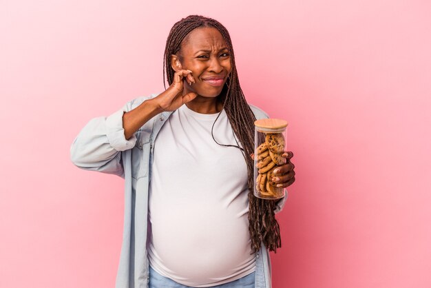 Giovane donna incinta afroamericana che tiene i biscotti isolati su sfondo rosa che copre le orecchie con le mani.