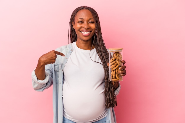 Giovane donna incinta afroamericana che tiene i biscotti isolati su sfondo rosa persona che indica a mano uno spazio copia camicia, orgoglioso e fiducioso