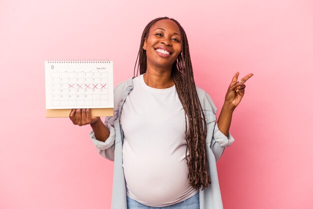 Giovane donna incinta afroamericana che tiene il calendario isolato su sfondo rosa gioioso e spensierato che mostra un simbolo di pace con le dita.