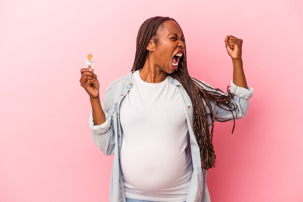 Giovane donna incinta afroamericana che tiene il ciuccio isolato su sfondo rosa alzando il pugno dopo una vittoria, concetto vincitore.