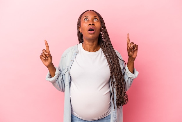 Giovane donna incinta afroamericana isolata su fondo rosa che indica al rialzo con la bocca aperta.