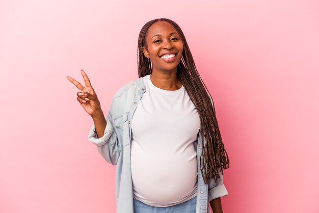 Giovane donna incinta afroamericana isolata su fondo rosa gioiosa e spensierata che mostra un simbolo di pace con le dita.