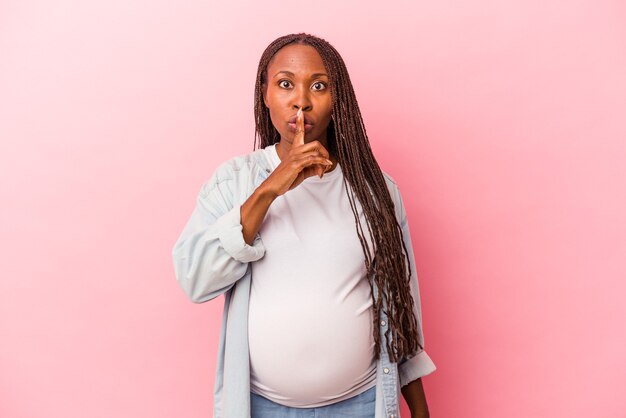 Giovane donna incinta afroamericana isolata su sfondo rosa mantenendo un segreto o chiedendo silenzio.