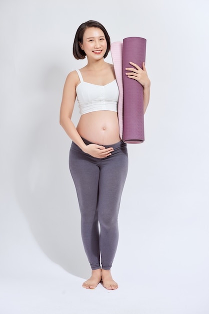 Giovane donna incinta attraente che tocca la sua pancia mentre tiene la stuoia di yoga nelle mani, gestante che fa esercizi fisici, posa isolata su sfondo bianco.