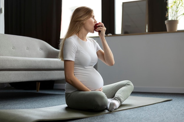 Giovane donna incinta che mangia mela rossa che pratica yoga seduto nella posa del loto Dieta yoga in gravidanza