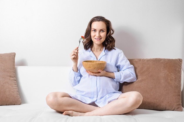 Giovane donna incinta che si siede sul sofà e che mangia insalata verde