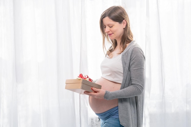 Giovane donna incinta con la grande pancia che tiene un regalo con la festa della doccia di bambino del nastro rosso con i regali