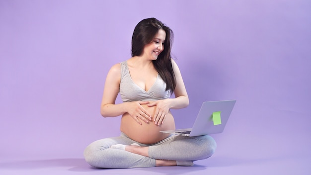 giovane donna incinta con una grande pancia pone in uno studio con un computer portatile su uno spazio viola. pancia nuda di una donna incinta.