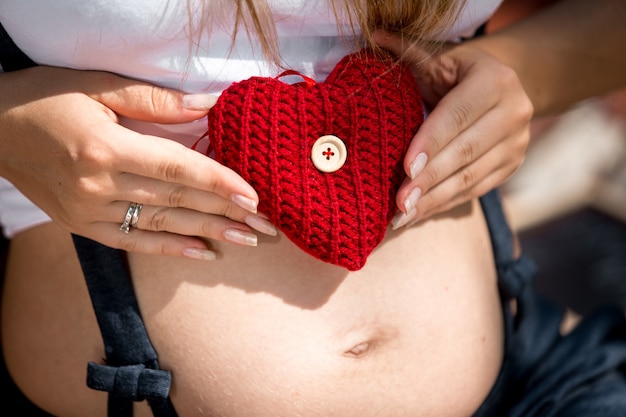 Giovane donna incinta del primo piano che tiene cuore rosso sull'addome