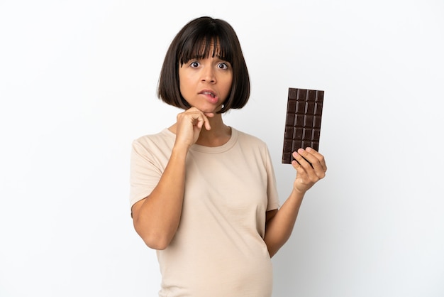 Giovane donna incinta di razza mista isolata su sfondo bianco prendendo una tavoletta di cioccolato e avendo dubbi