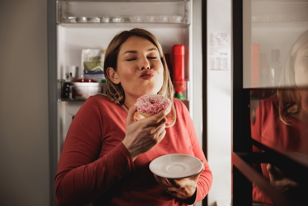 Giovane donna incinta felice che mangia ciambella davanti al frigorifero aperto in cucina di notte.