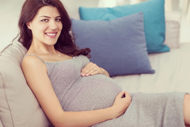 giovane donna incinta felice che tiene la mano sulla pancia mentre è seduta sul divano a casa