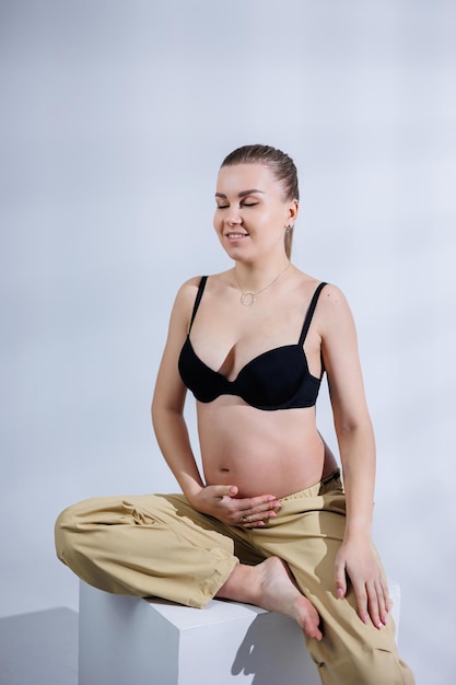 Giovane donna incinta felice in reggiseno e pantaloni su sfondo bianco Gravidanza felice Elegante donna incinta pone in studio