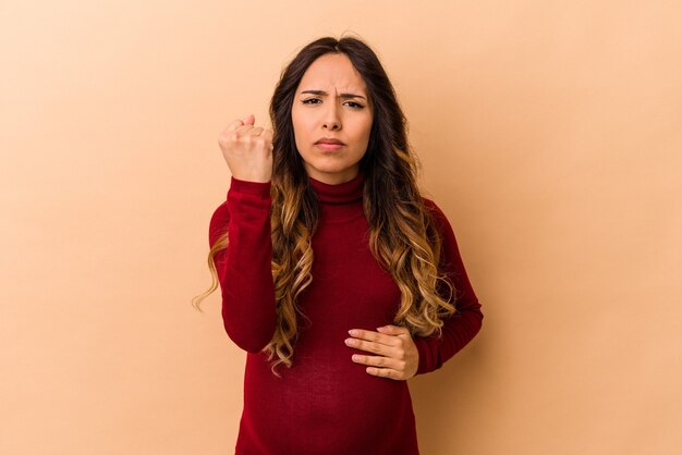 Giovane donna incinta messicana isolata su fondo beige che mostra pugno alla macchina fotografica, espressione facciale aggressiva.