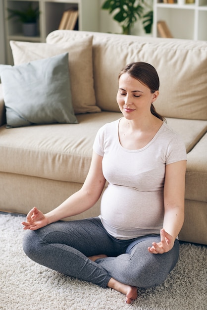 Giovane donna incinta positiva in abbigliamento sportivo seduto nella posa del loto sul divano sul pavimento e meditando mentre si resta a casa